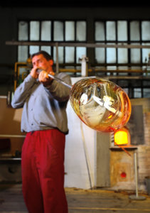 Man blows glass on end of long pole in the Moser glassworks factory