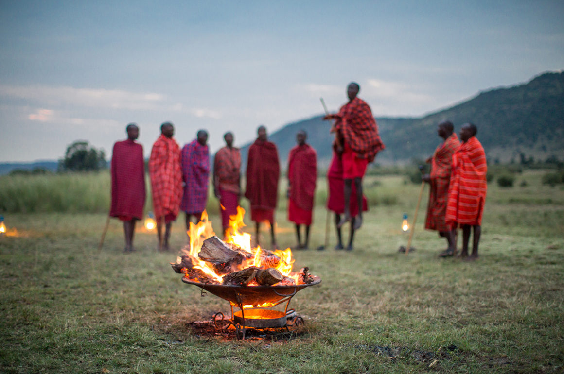 Massai warriors in red traditional dress jumping behind a fire in the African bush