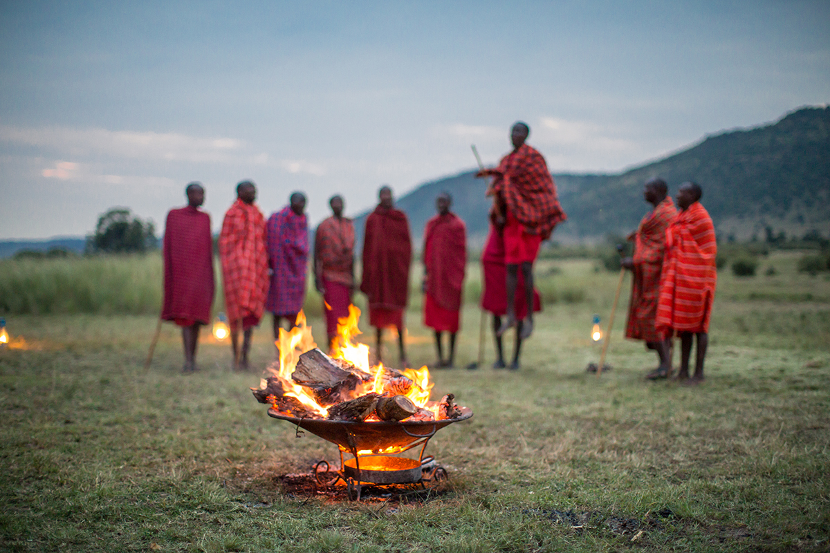 Massai warriors in red traditional dress jumping behind a fire in the African bush