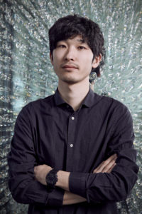 Japanese man wearing navy blue shirt standing in front of crystal background with arms folded