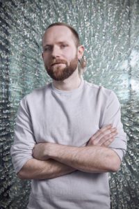 Bearded man standing in front of wall of crystals with arms crossed