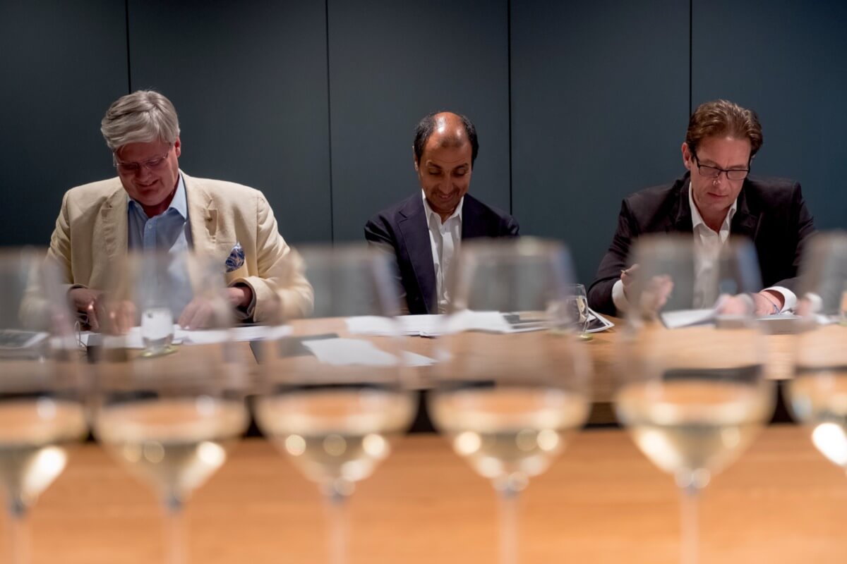 line of wine glasses with three men in suits sitting behind considering documents