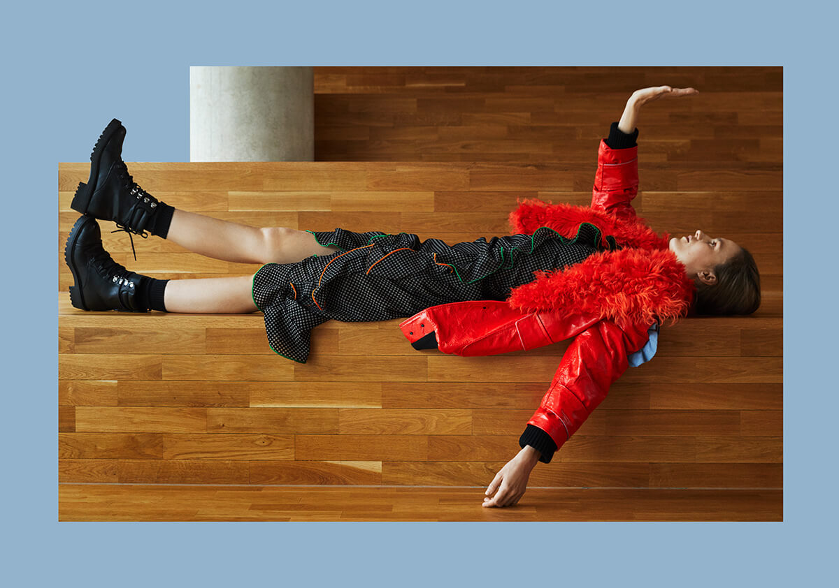 woman lying on her back wearing black dress, boots and red jacket with one arm lifted to the ceiling