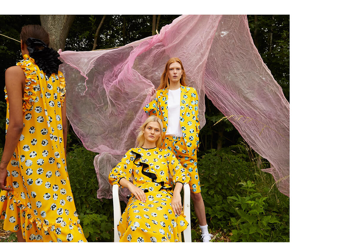 Three female models pose in front of billowing pastel pink fabric wearing yellow patterned dresses and coats