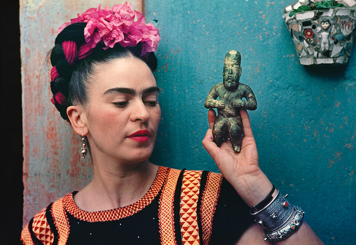 Colour photograph of Mexican artist Frida Kahlo holding a traditional figurine against a blue door