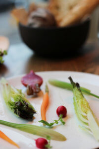 plate of artistically arranged baby vegetables on a white plate with bread basket in background