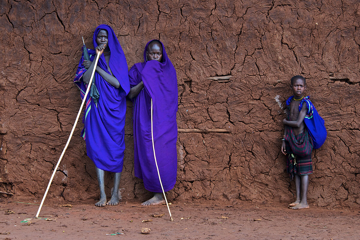 Omo valley tribesmen dressed in bright blue cloth holding wooden sticks and standing against a red mud wall