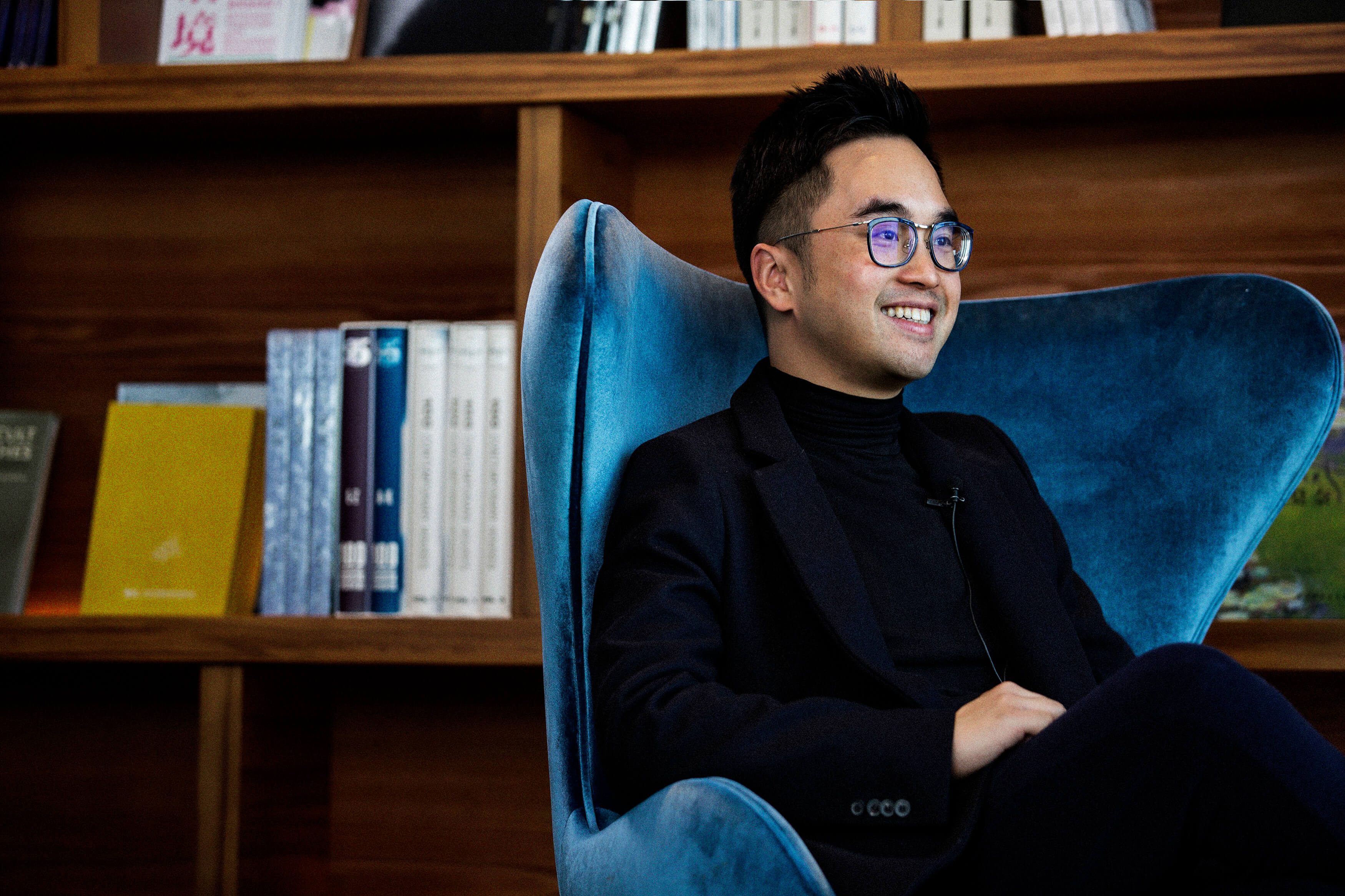 Man wearing black polo neck sitting on blue velvet chair wearing glasses with wooden bookshelves behind