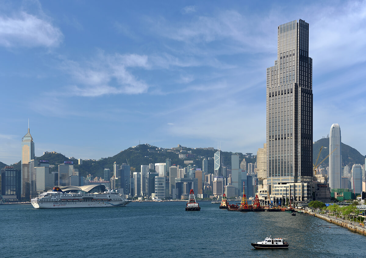 Tall skyscraper on the edge of waterfront with boats floating, buildings and mountains in the distance 