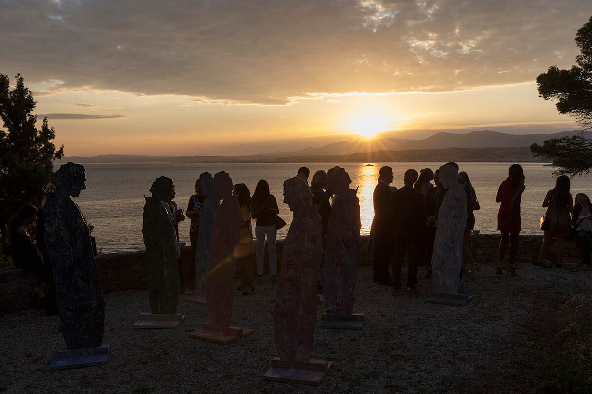 A crowd of people and sculptures on the cliff edge as the sun sets over the ocean in the distance