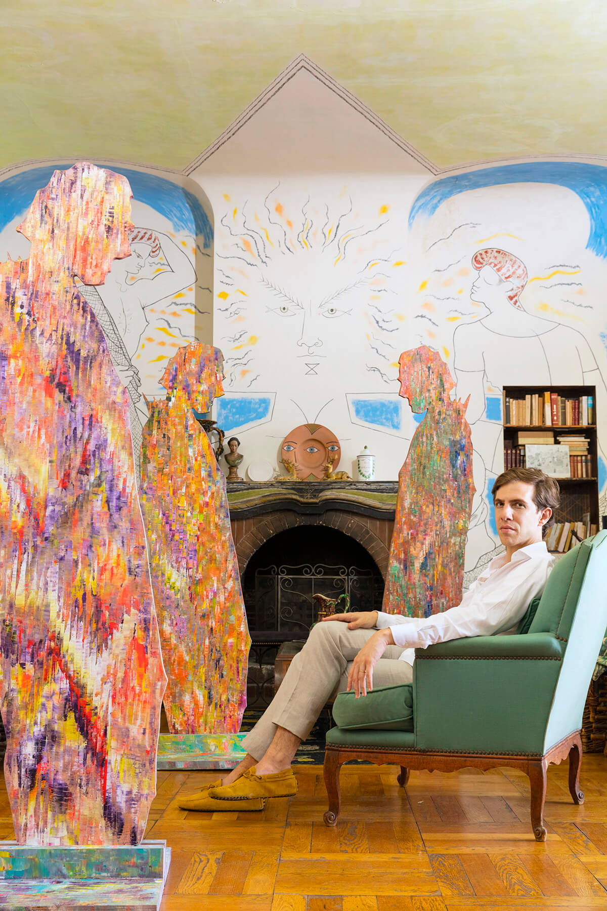 artist Sassan Behnam-Bakhtiar seated in a green arm chair surrounded by colourful sculptures of men and a painted mural