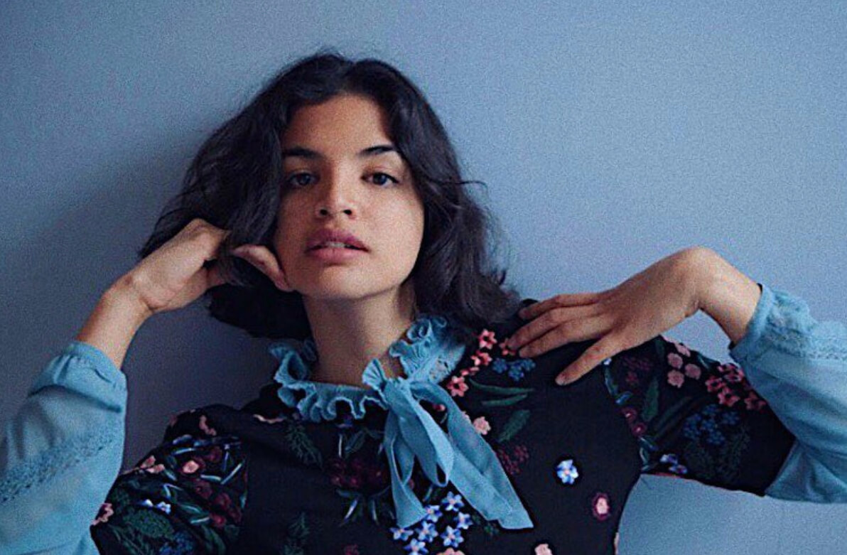 Model poses against pale blue wall wearing ruffled collar shirt with short black hair