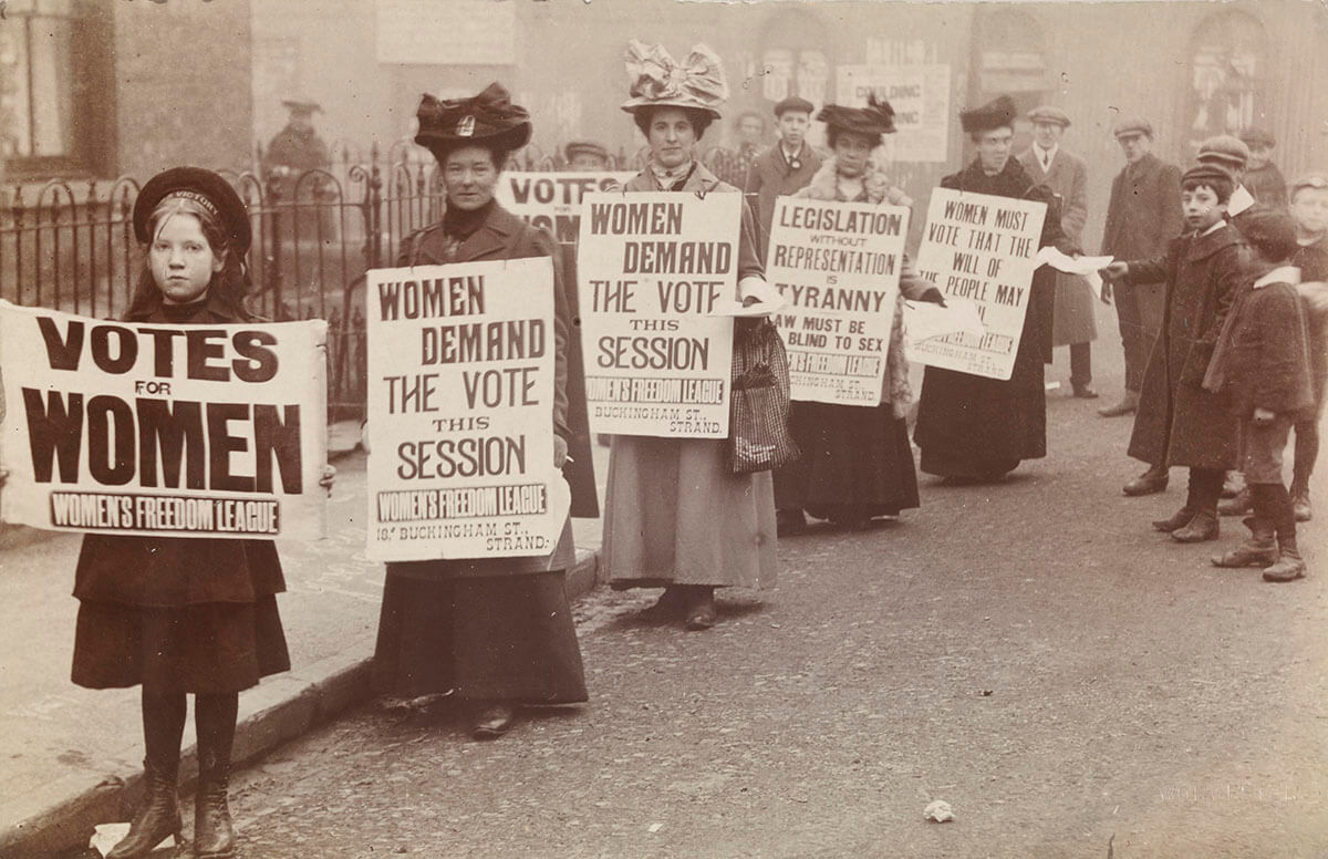Historical suffragette photograph in black and white of women's parade holding signs with the suffrage message