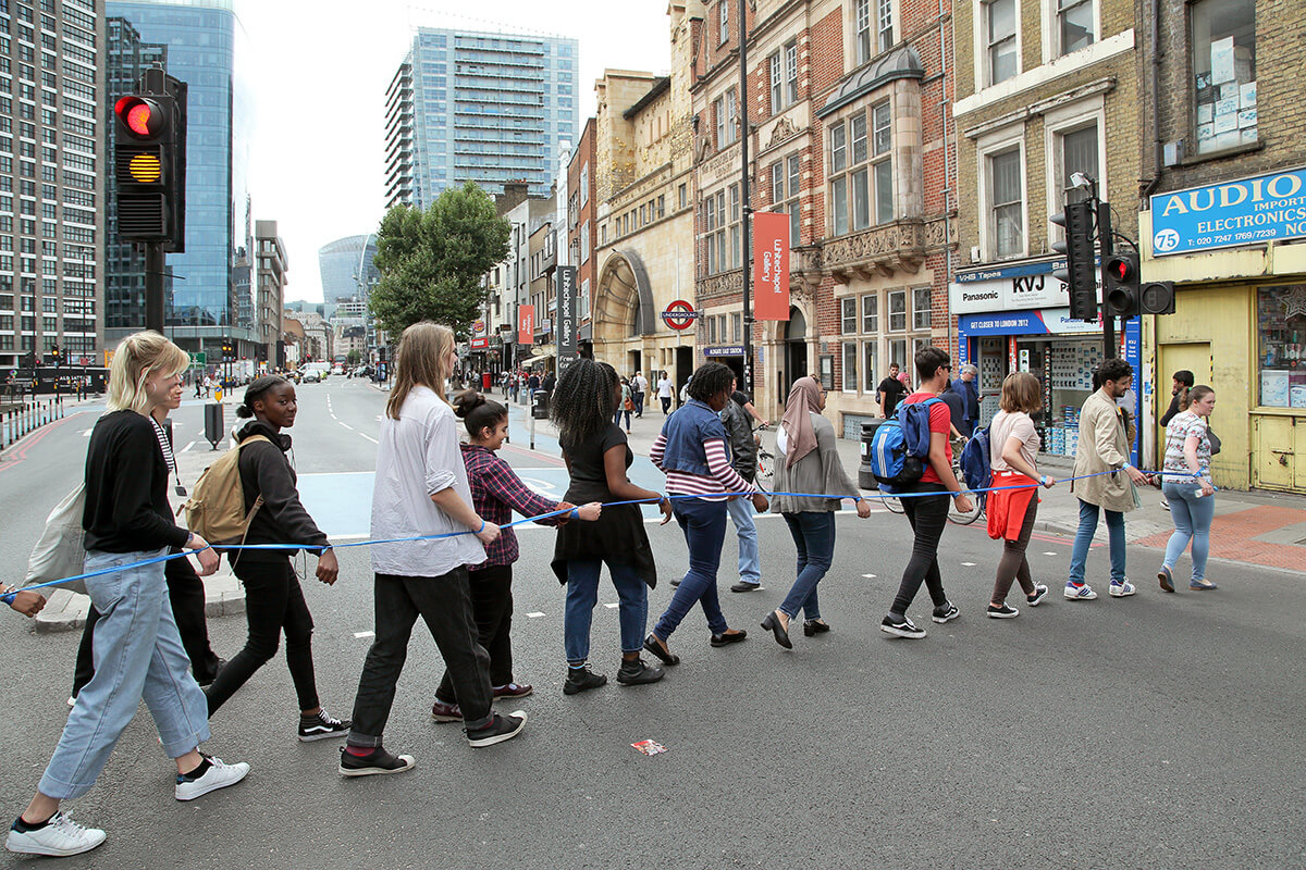 A live street art performance featuring people walking along a blue rope across a road