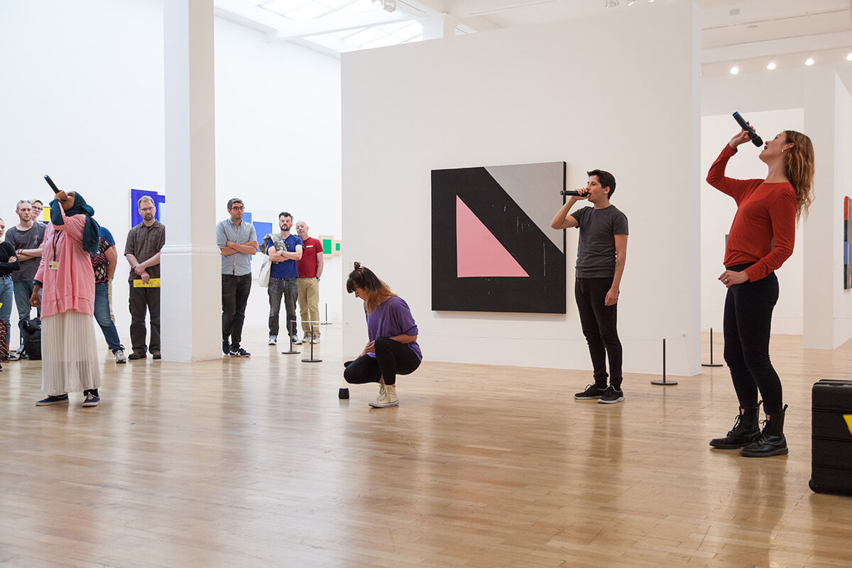 Performance art piece featuring a man speaking into a microphone and a woman kneeling in gallery setting