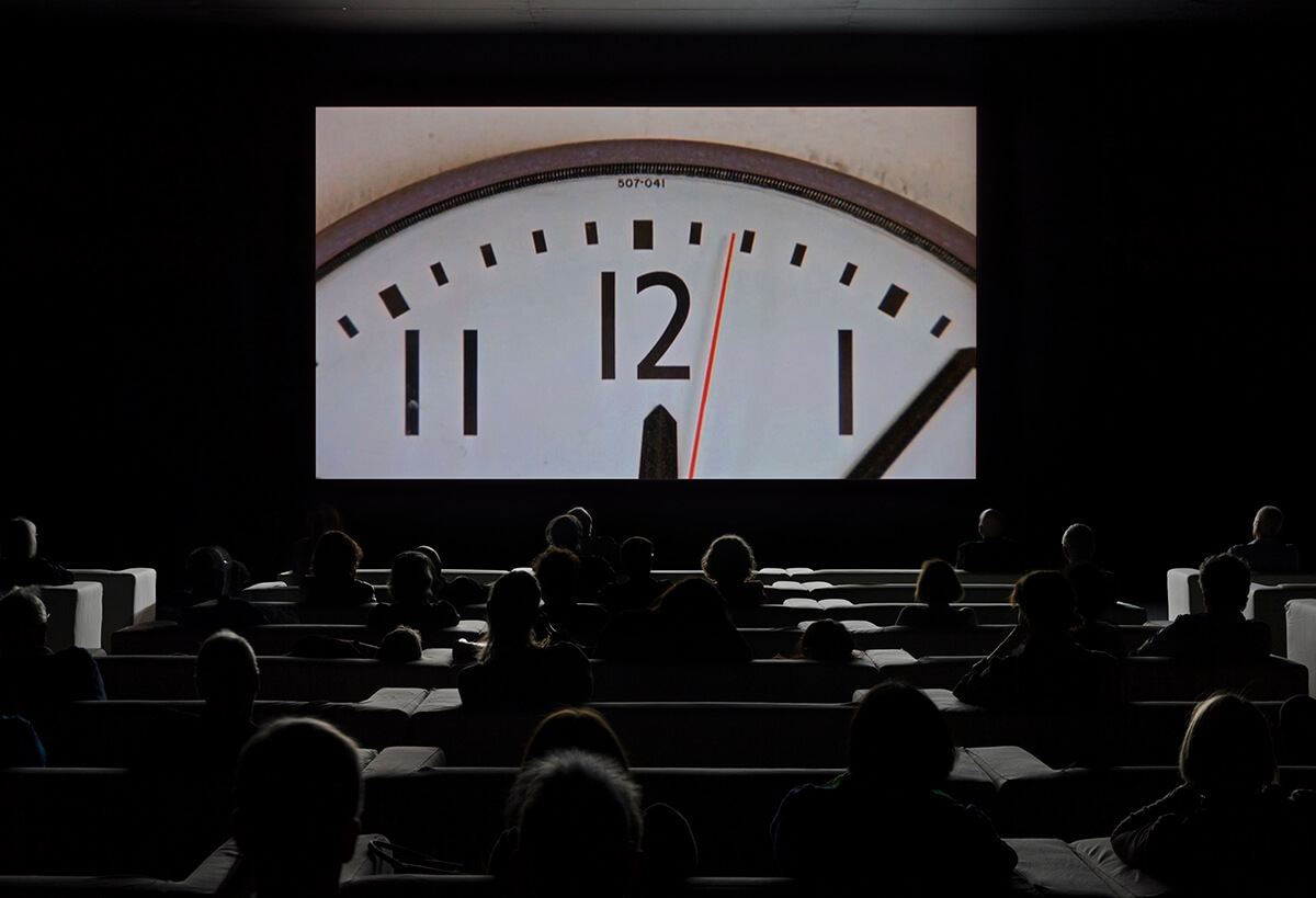 Cinema with a screen showing the top of a clock with people sitting in rows of seats 