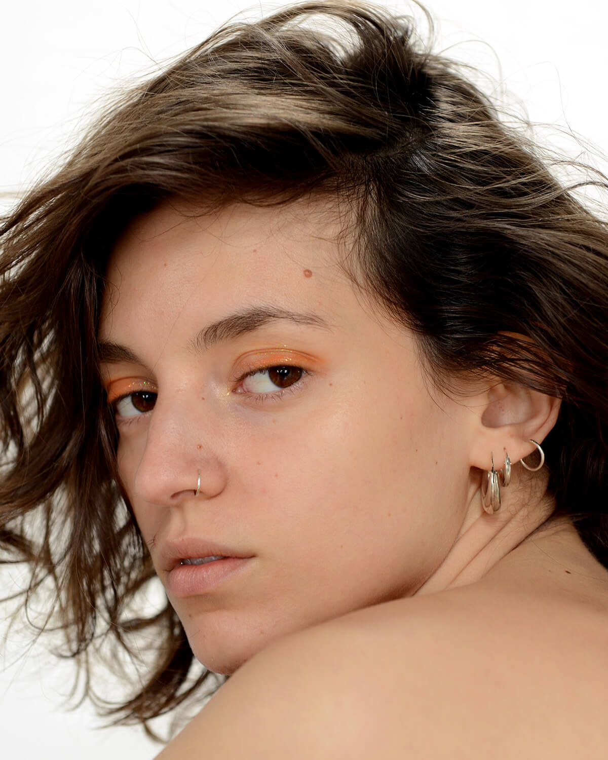 Portrait of a young woman with short brown hair looking over a bare shoulder wearing orange eye shadow