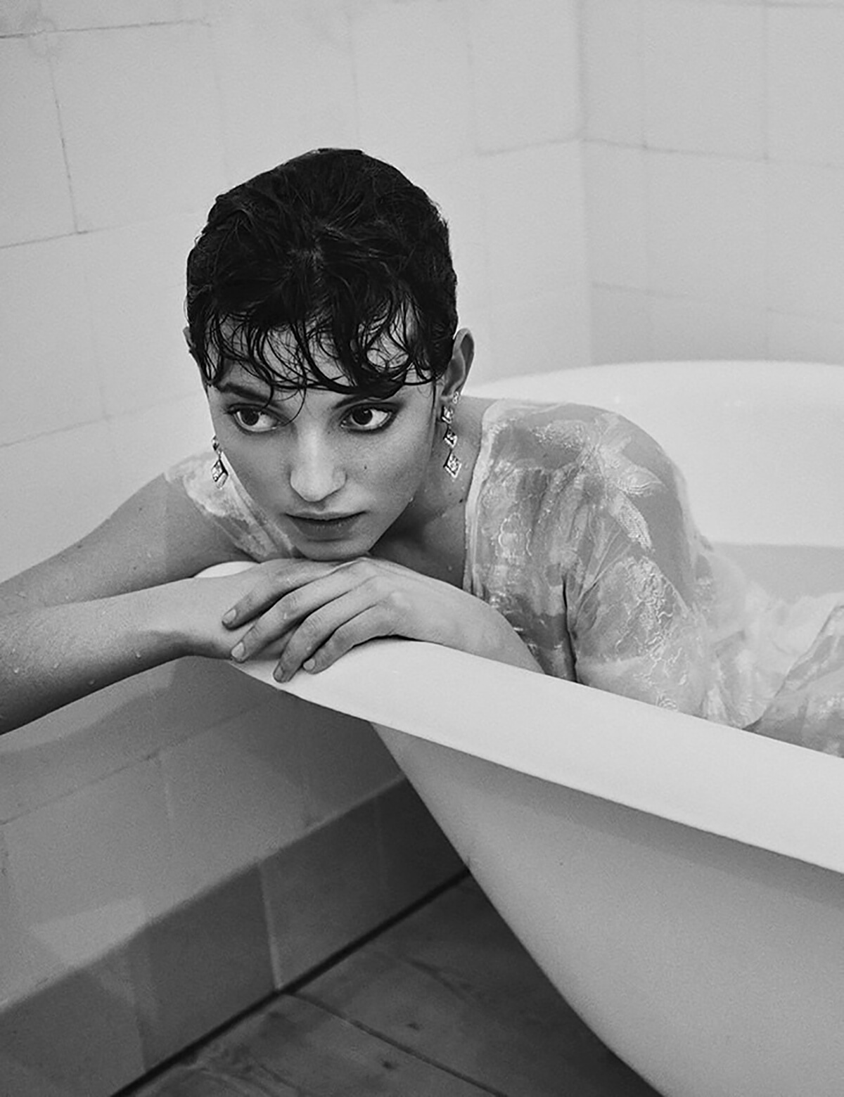 Black and white image of a woman in a bath wearing a white tshirt with dark hair and make-up resting her head on her hands on the bathtub rim