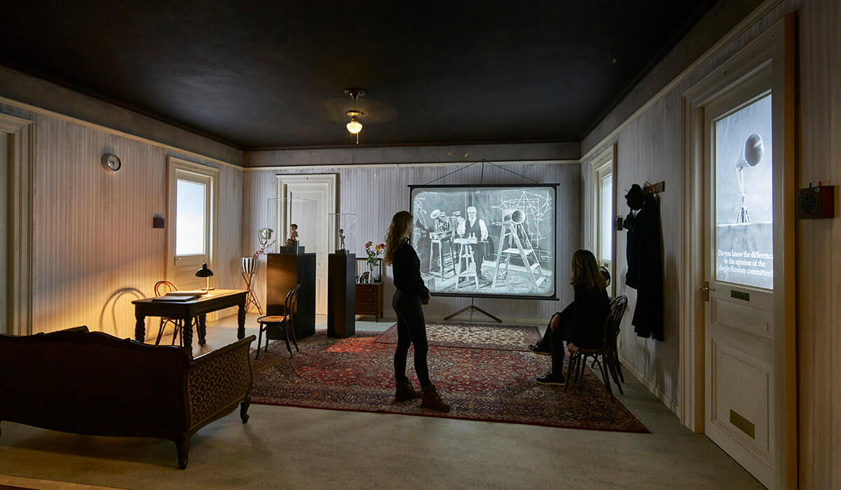 Multimedia art installation with screen showing black and white film and living room set up