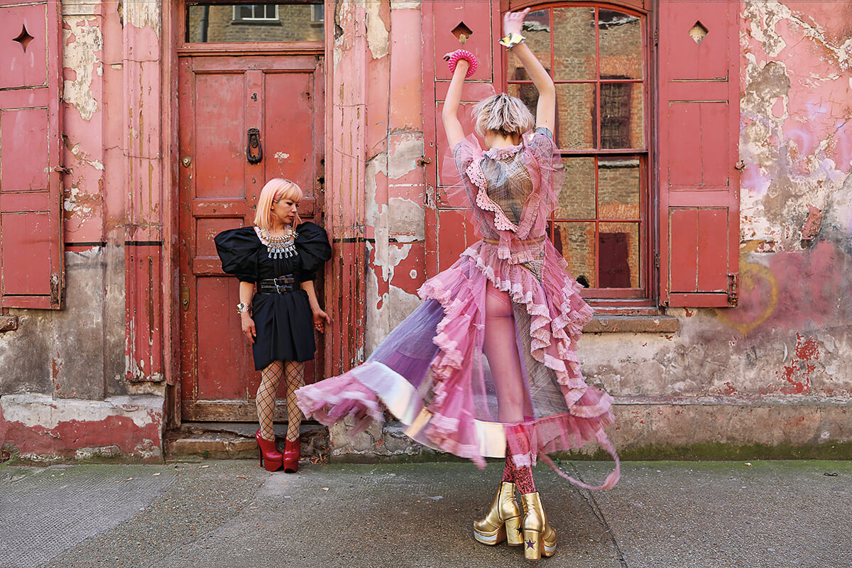 Model poses on street in mid dance in pink frilly dress with designer in background