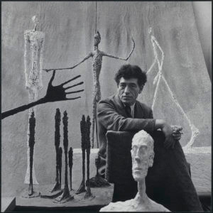 Black and white portrait of Alberto Giacometti in his studio surrounded by sculptures