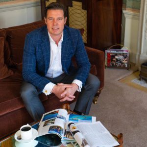 Portrait of a man wearing a suit sitting on a sofa with magazines open on a table in front of him