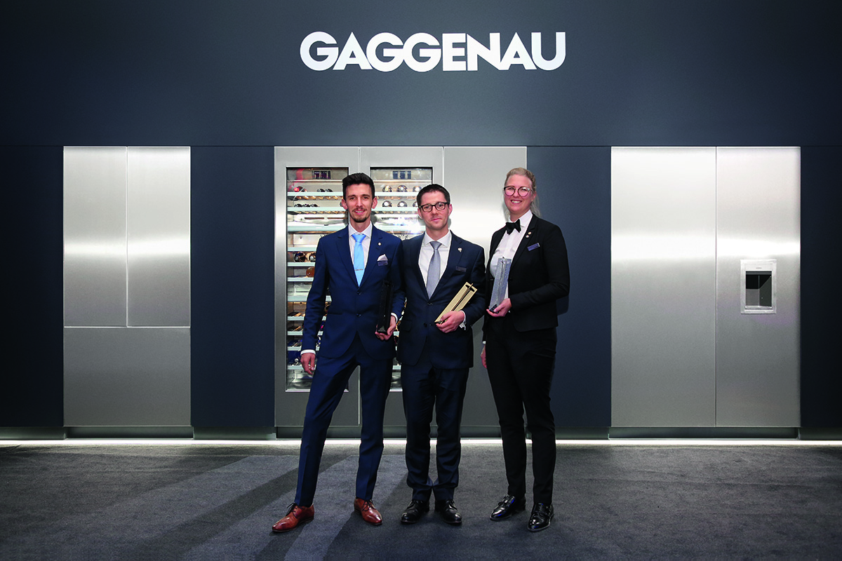 Two men and a woman stand in front of Gaggenau sign and sleek metal cabinet
