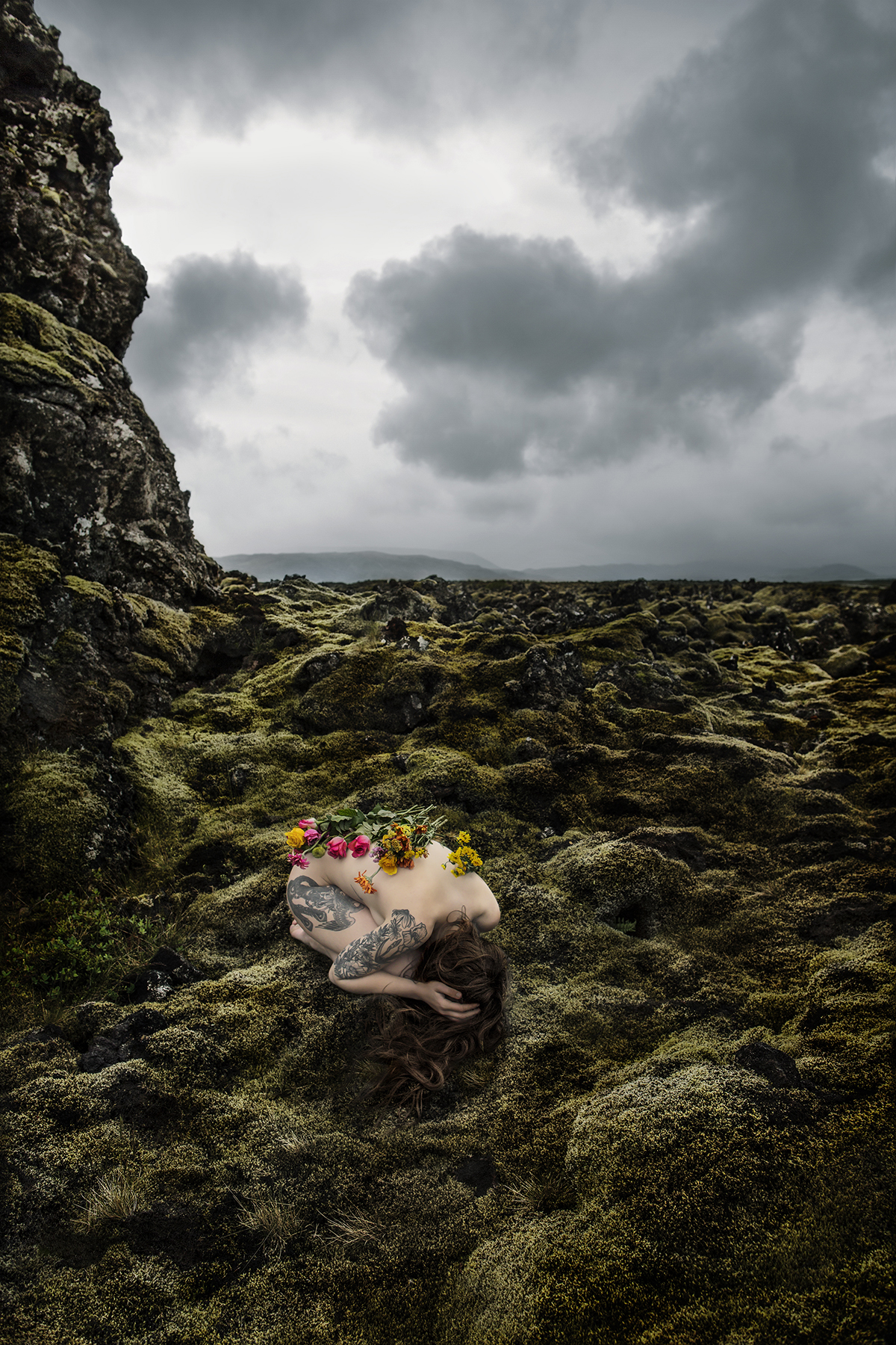 A naked woman crouched in a green bare landscape with flowers on her spine 