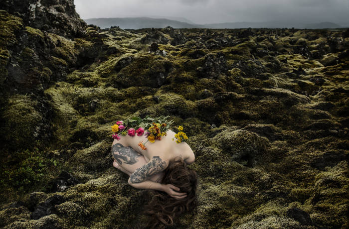 A naked woman crouched in a green bare landscape with flowers on her spine