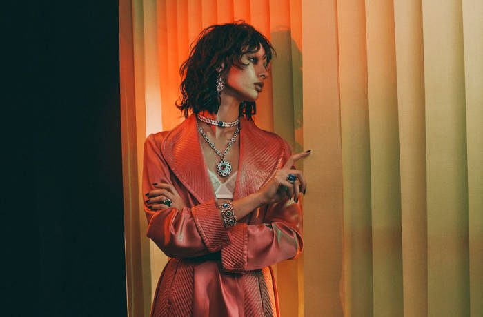 Model stands looking out of blinds wearing multiple jewels