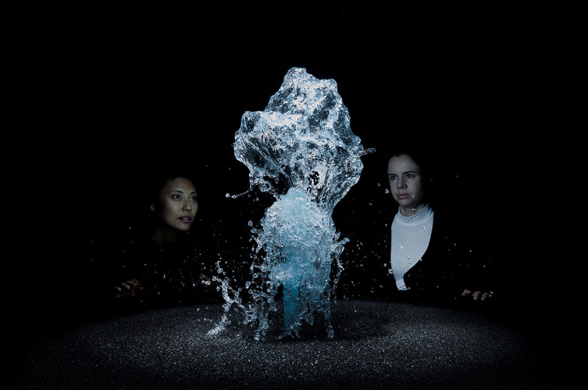 Two women watch water rising up out of a surface in a black room
