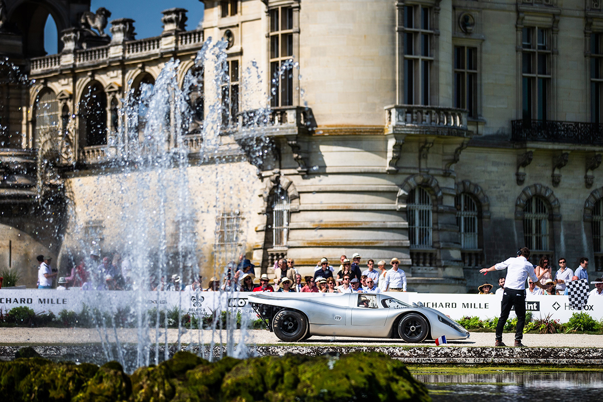 A silver sports car pictured in front of a stately home and behind a water fountain
