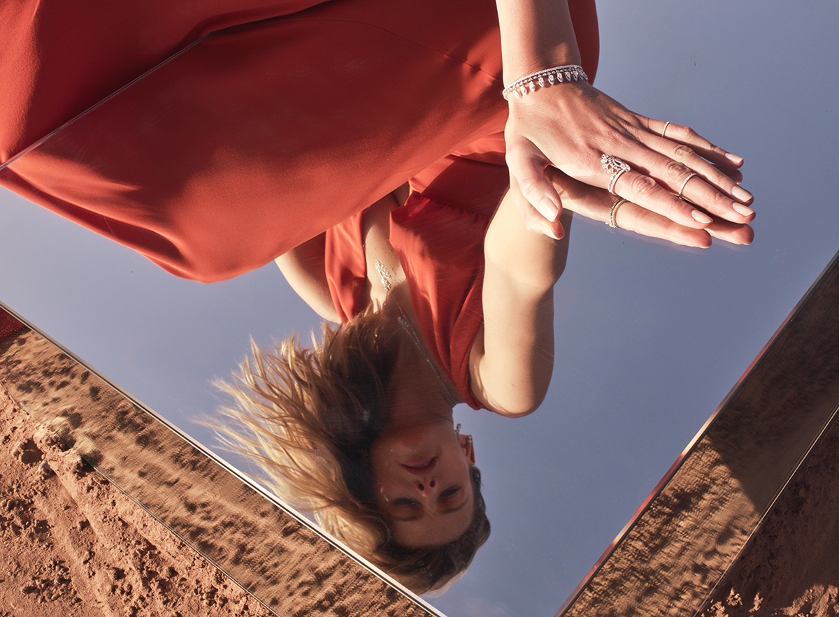 Model leaning over a mirror wearing a red dress and diamond jewellery