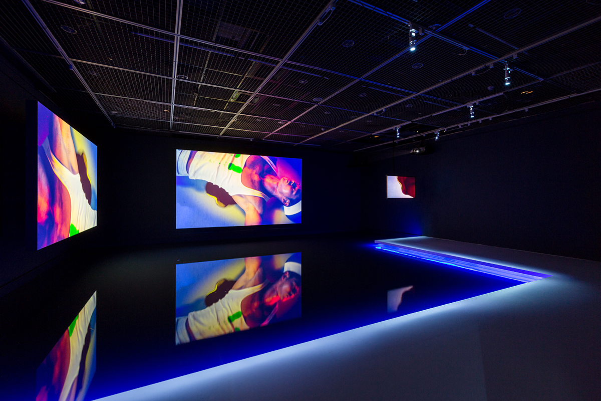 three screens with coloured lights in a dark room showing a man lying down wearing a vest