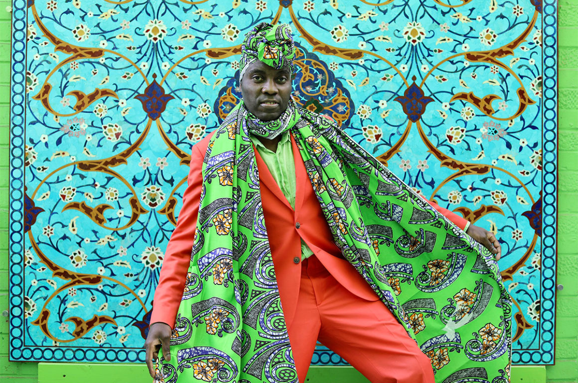 man in an orange suit and green patterned scarf and hat standing in front of a patterned wall