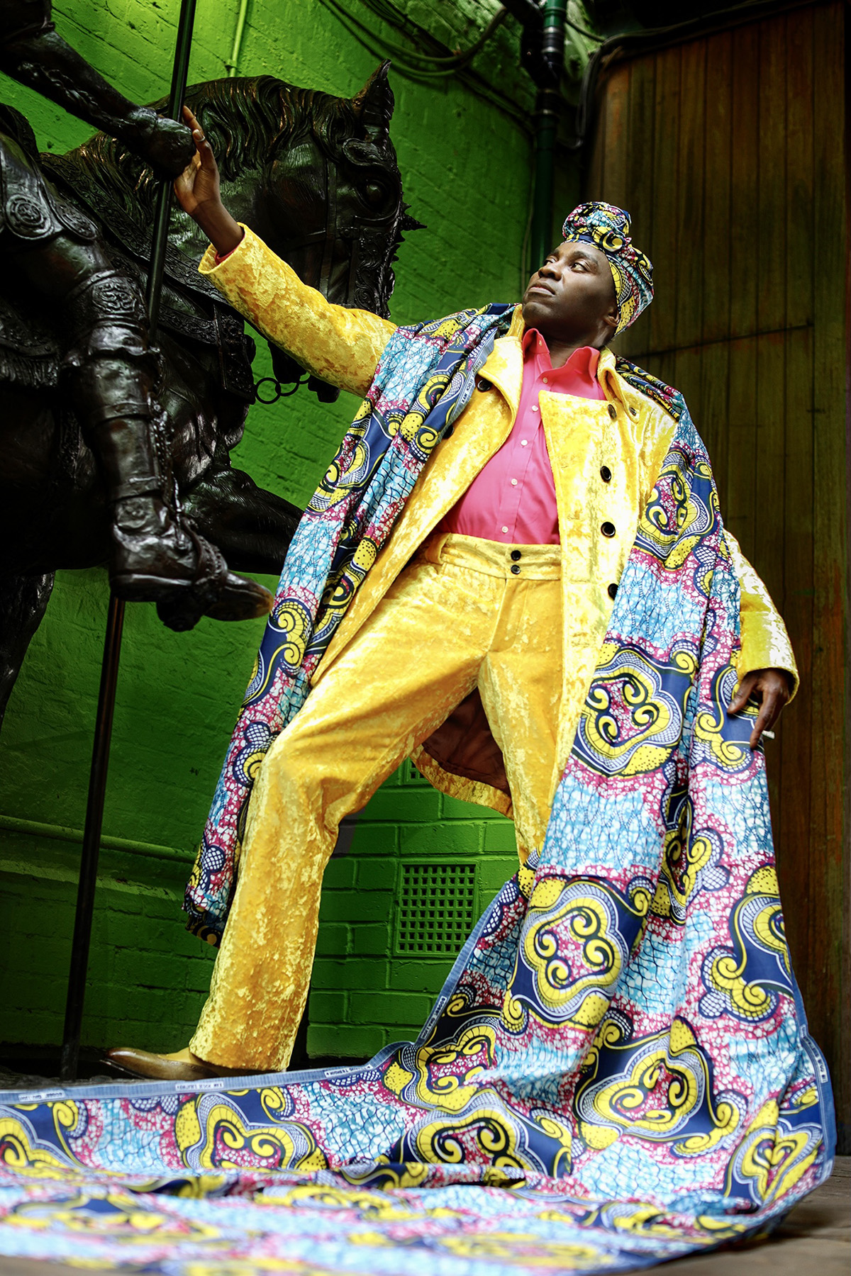 A man in a yellow suit standing by a green wall wearing a long colourful scarf