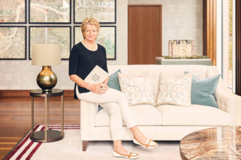 Penny Hughes wearing a black top and white trousers holding a book sitting on the arm of a sofa