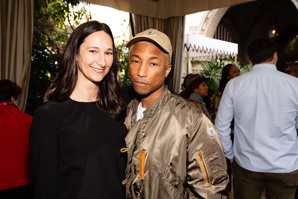 A woman in a balck top standing next to Pharrell Williams in a big kaki puffer coat and cap