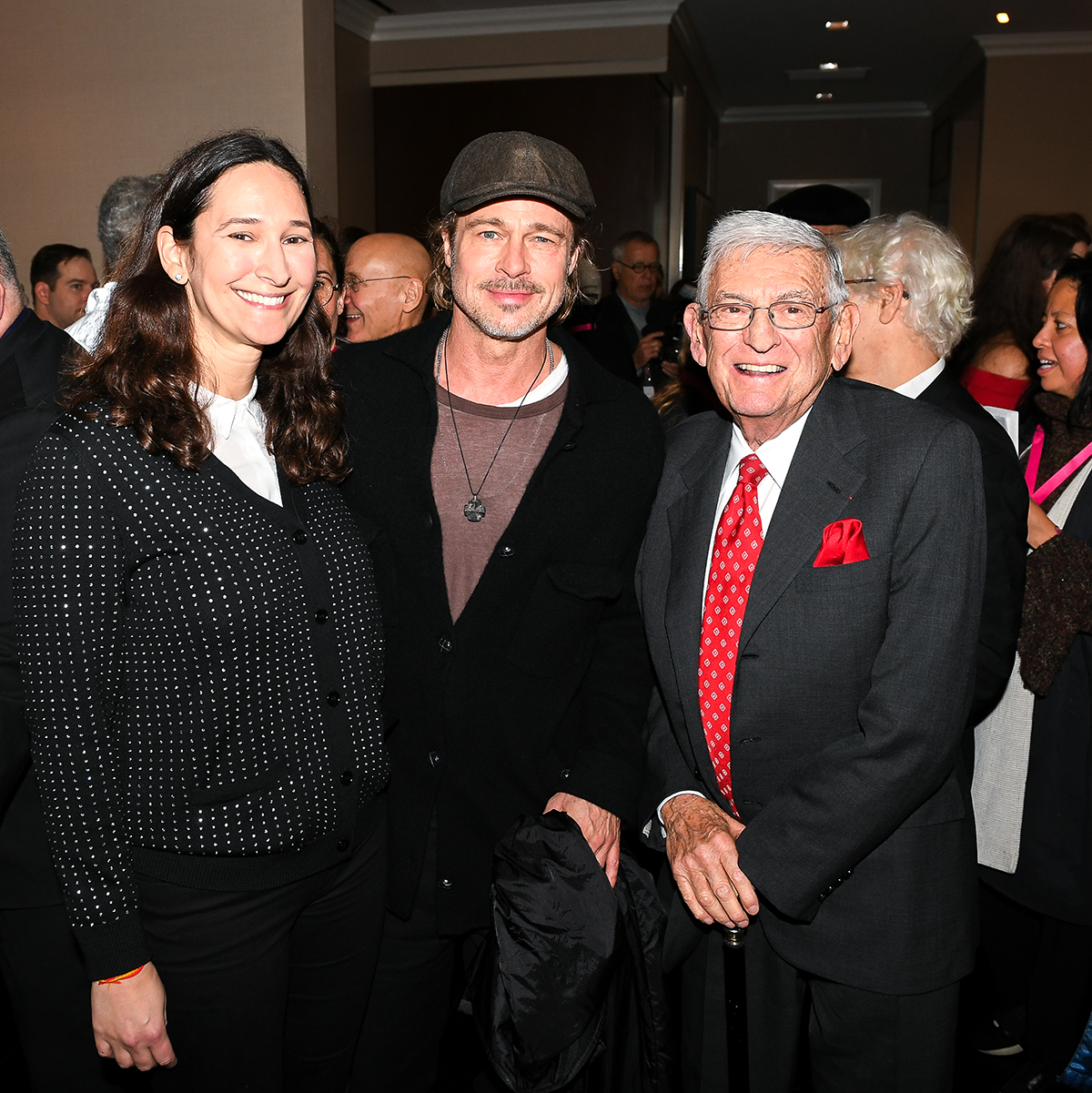 A woman standing next to Brad Pitt, in a hat jumper and coat and another man wearing a red tie and pocket handkerchief
