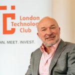 grey suit and pink shirt sitting in front of a sign that says 'London Technology Club'