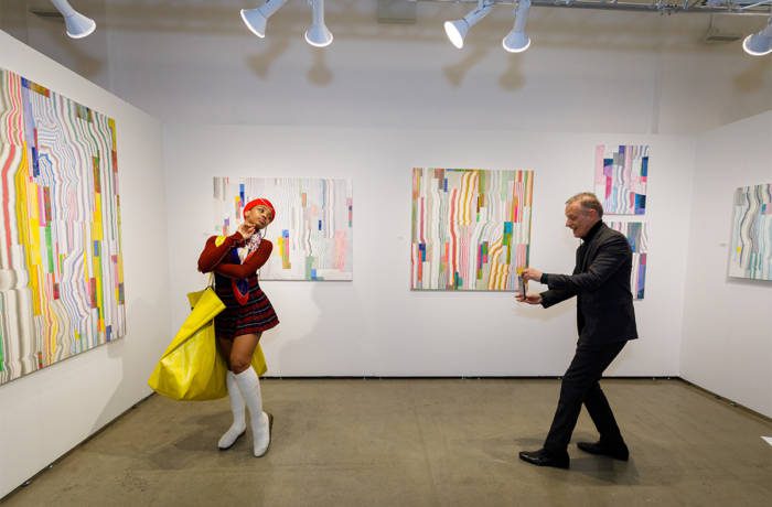 A girl posing in an art gallery and a man taking a photo on his IPhone