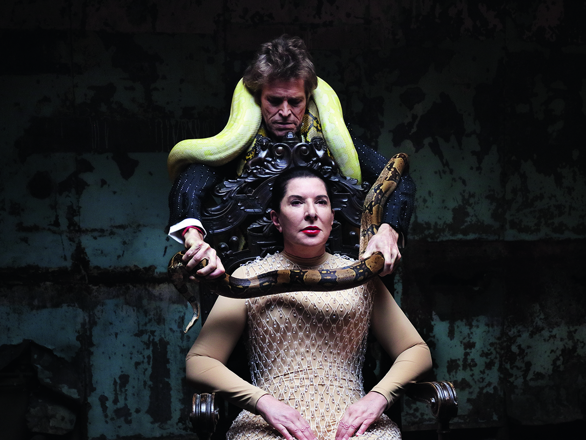 A man with a yellow snake wrapping a brown snake around a woman on a bedazzled top sitting on a chair