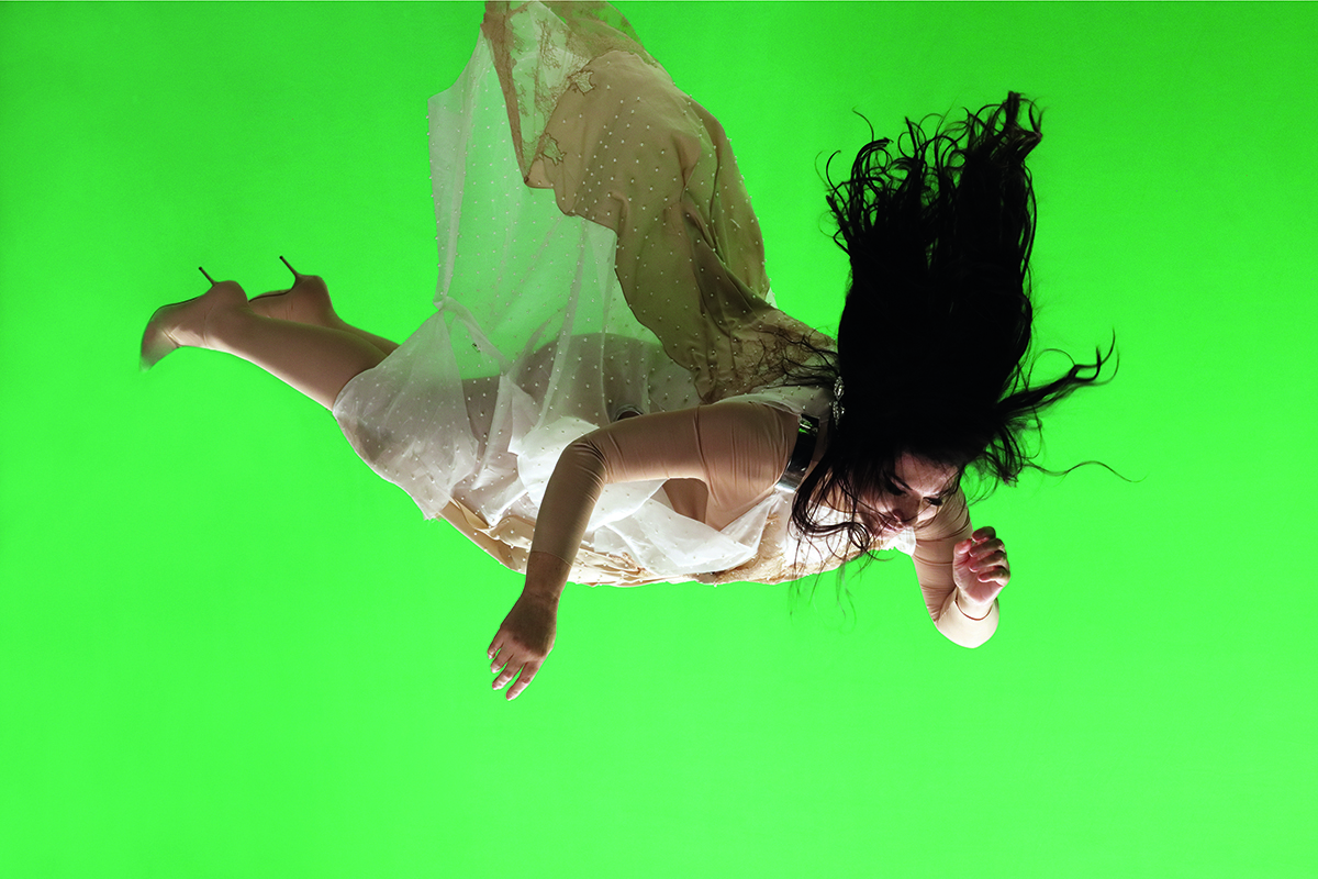 A woman falling through the air with a green background wearing a nude coloured dress and heels