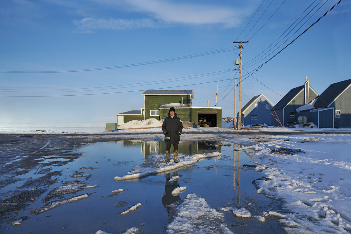 Jordin Tootoo standing on a piece of ice in the sea with a power line and green house behind him