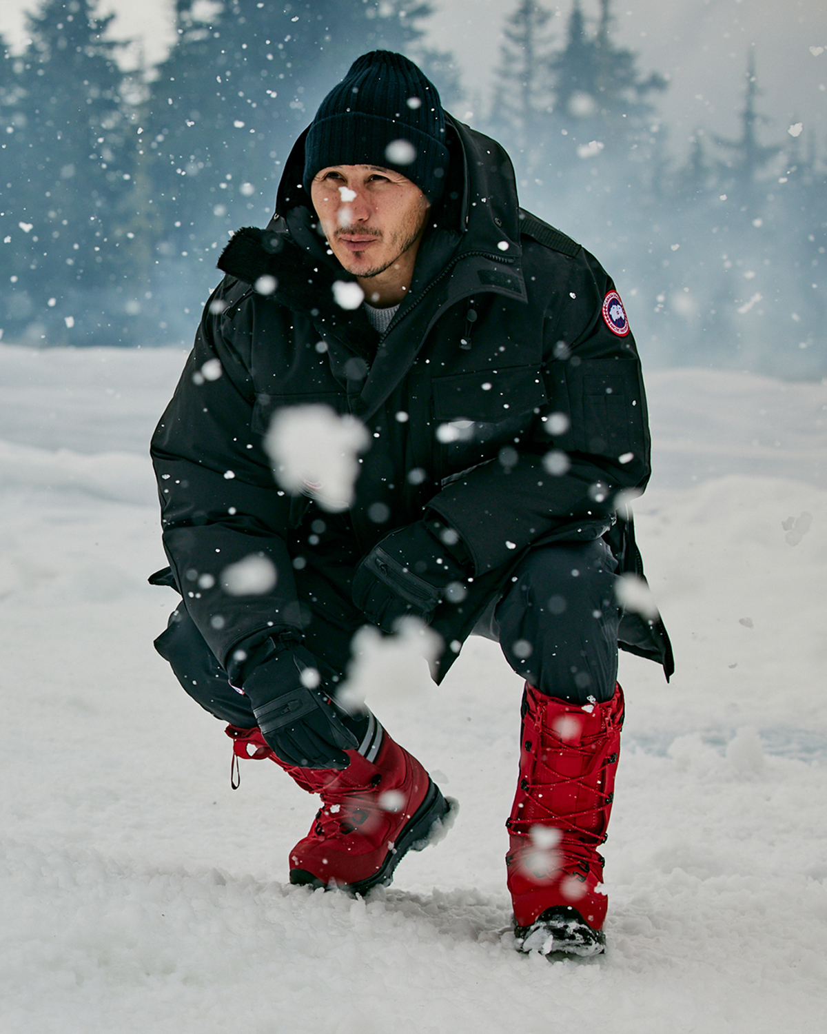 A man in a black coat and red boots squating over the snow