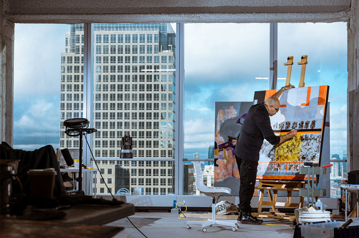 A man painting by a window with a skyscraper outside