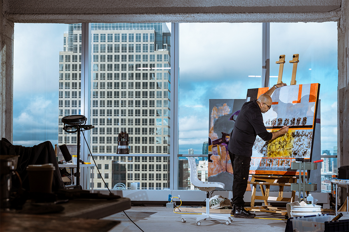 A man painting by a window with a skyscraper outside