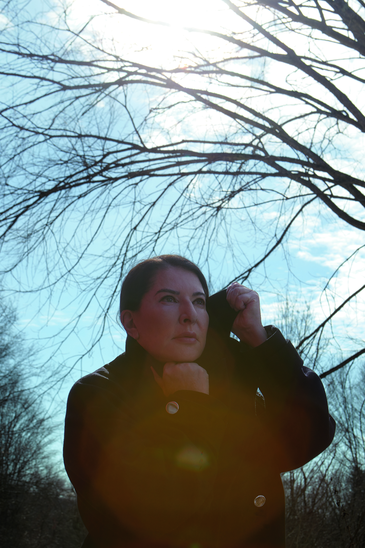 A woman outside by a tree with clouds in the sky wearing a black coat