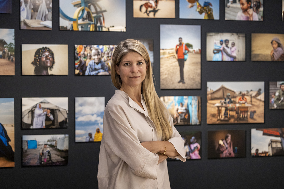 A blonde woman in a beige top standing in front of a wall of photographs with her arms crossed