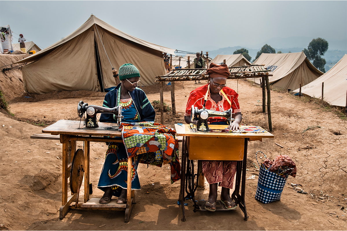 Two women sewing clothes with a tent behind them